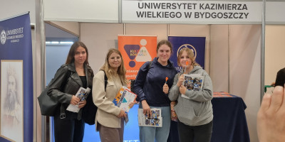 Cztery maturzystki stoją przed stoiskiem Uniwersytetu Kazimierza Wielkiego w Bydgoszczy, uśmiechają się