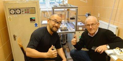 Kei Kamada i Yuriy Zorenko pozują do zdjęcia, pokazując wyciągnięte kciuki. Za nimi sprzęt laboratoryjnyy